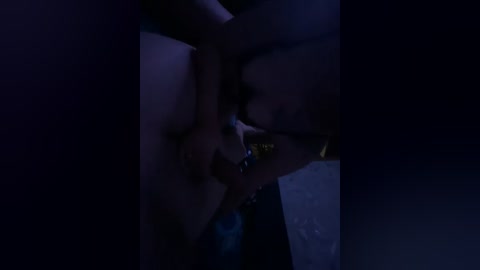 Video of a dimly lit, intimate moment between two people. A woman with fair skin and long, dark hair is passionately kissing a man with a trimmed beard and glasses. The background is dark and blurry, enhancing the sensuality of the scene.