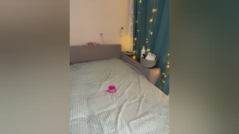 Video of a cozy bedroom with a gray bedspread, a pink toy car on the bed, teal curtains with string lights, a bedside table with a lamp and a bowl, and a cream-colored wall.