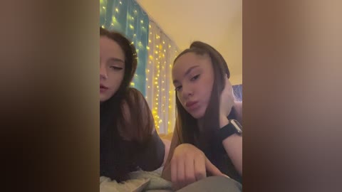 Video of two young women with fair skin and long brown hair, one wearing a black top, seated closely together, in a cozy room with yellow fairy lights and a plush bed.