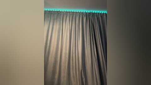 Video of a gray curtain with a subtle green LED strip along the top, creating a soft, ambient glow. The curtain is evenly pleated, and the background is a plain, light beige wall.