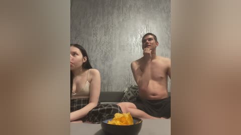 Video of a man and woman, shirtless and wearing plaid pajama pants, sitting on a couch. The man holds a bowl of popcorn, while the woman has a neutral expression.