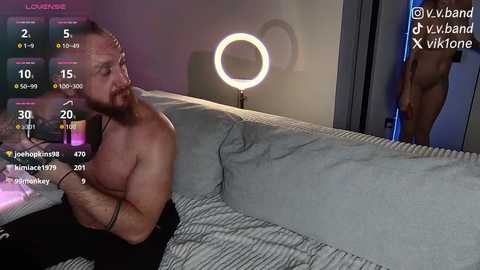 Video of a shirtless, bearded man sitting on a gray couch, playing a video game with a virtual band performing in the background.