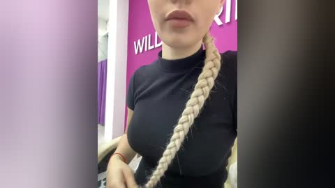 A video of a woman with fair skin, wearing a black turtleneck, and a long, braided blonde hair accessory. The background features a purple wall with the word \"WILL\" partially visible.