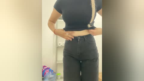 Video of a woman with light skin and medium build wearing a black cropped top and high-waisted black jeans, standing in a bright, minimalist room with a white wall and a blue water bottle on the floor.
