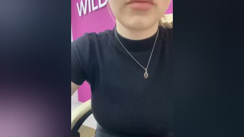 Video of a woman in a black t-shirt, seated, wearing a silver necklace with a pendant, in a room with a pink wall and the word \"WILD\" partially visible.