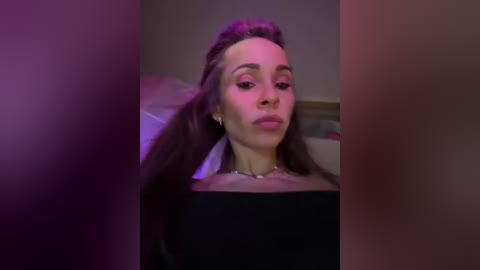 A video of a young woman with long, dark hair styled in a voluminous updo, wearing a black top and minimal makeup. She's in a dimly lit room with blurred, purple-hued lighting.