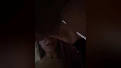 A dimly-lit video shows a close-up of a woman's face with long dark hair, partially obscured by a man's hand. The background is dark, creating a mysterious and intimate atmosphere.