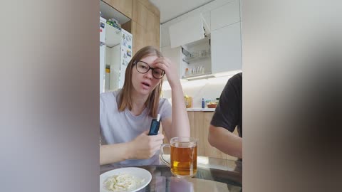 Video of a woman with glasses and straight light brown hair, wearing a light blue t-shirt, sitting at a glass-top table, holding a blue phone, looking tired. Background features a modern kitchen with wooden cabinets, a white fridge, and a glass of beer.