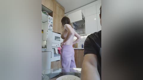 A candid video shows a topless woman in light pink pants adjusting her clothing in a modern kitchen with white cabinets, wooden accents, and a stainless steel fridge.