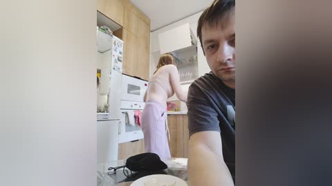 A video depicts a light-skinned man in a grey t-shirt and a woman in a pink towel, cooking in a modern kitchen with wooden cabinets.