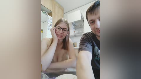 Video of a light-skinned woman with glasses and straight brown hair, topless, smiling at a man with short dark hair in a kitchen.