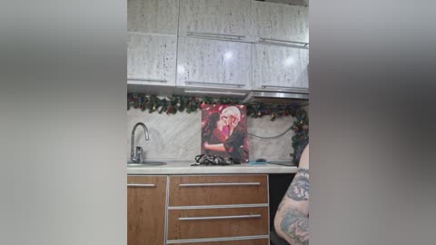 A video of a modern kitchen featuring light wooden cabinets, white marble backsplash, a framed Marilyn Monroe poster, and a tattooed arm.