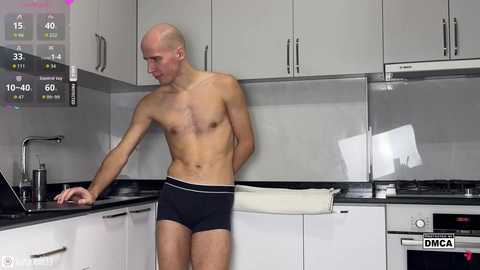 Video of a shirtless, muscular, bald man in black underwear, standing in a sleek, modern kitchen with white cabinets and black countertops.