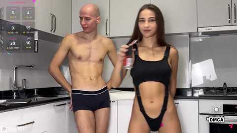 Video of a fit, bald man wearing black boxer briefs and a slender, brunette woman in a revealing black one-piece swimsuit, standing in a modern kitchen with white cabinets.