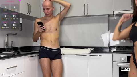 Video of a shirtless, slim, bald Caucasian man in navy boxer briefs, holding a smartphone, in a modern white kitchen. Another person, partially visible, is handing him a white towel.