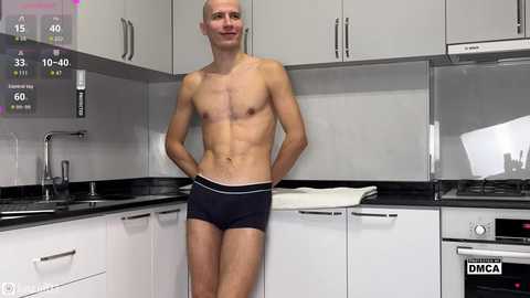 Video of a shirtless, fit, bald man wearing black briefs standing in a modern, white kitchen with black countertops, a sink, and digital display showing 15\u00b0C.