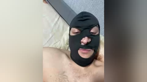 Video of a topless man with a black hood covering his face, lying on a bed with beige sheets, surrounded by a grey textured headboard.
