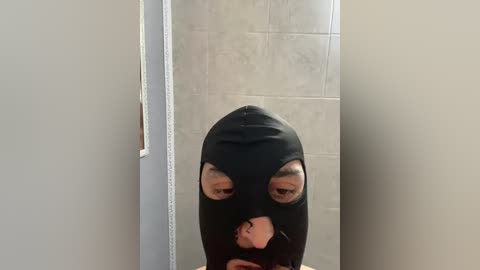 A video of a person in a black hooded mask with eye holes, standing in a tiled hallway. The mask obscures most of their face, and the background is plain and neutral.