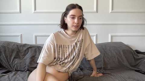 A video of a young woman with short dark hair, wearing a loose beige T-shirt and gray blanket, sitting on a gray quilted bed against a white paneled wall.