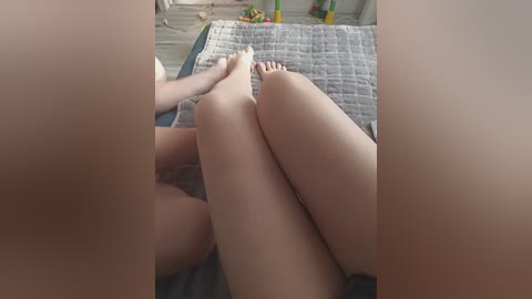 Video of a woman lying on her back, legs crossed, on a bed with a gray quilt, blurred hands holding her feet, and a green toy in the background.