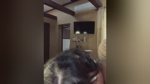 Video of a dimly lit bedroom with beige walls, dark wooden ceiling beams, a flat-screen TV mounted on the wall, and a partially visible person lying on a bed with a messy, dark hairdo.