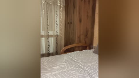A video of a cozy bedroom with a white bedspread, wooden headboard, and beige walls. A sheer curtain with a floral pattern covers a window, and a wooden door is partially visible on the right.