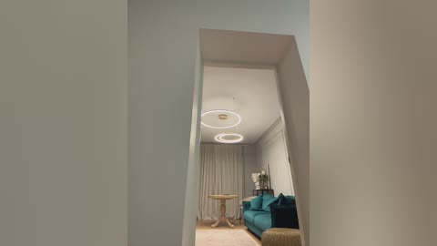 Video of a modern, minimalist living room with a teal sofa, white walls, a round wooden table, and two circular chandeliers.