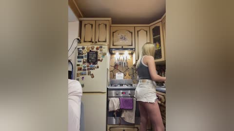 A video of a blonde woman in a gray tank top and high-waisted shorts cooking in a cozy, vintage-style kitchen with wooden cabinets, a white fridge covered in magnets, and a small oven.