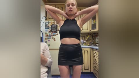 Video of a blonde woman in a black crop top and skirt, stretching in a cluttered kitchen with beige cabinets, a fridge covered in photos, and a person in the background.