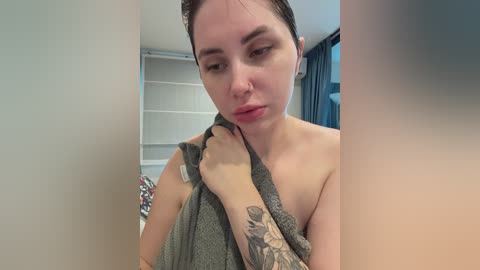 Video of a pale-skinned woman with dark hair, wearing a gray towel, holding it up to her neck, showing a tattoo sleeve on her left arm. Background includes a window with dark curtains and a bed with colorful bedding.