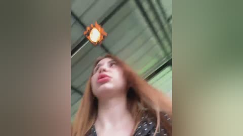 A blurry video of a young woman with fair skin, long red hair, and pink lipstick, standing under a sunlit industrial ceiling with exposed beams and a circular light fixture.