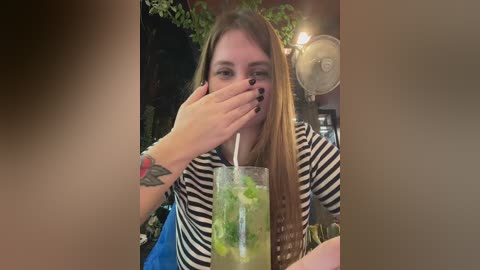 Video of a young woman with long brown hair, wearing a striped shirt, covering her mouth with her hand, sitting at a restaurant table, drinking a green drink with a straw, surrounded by greenery and a fan.