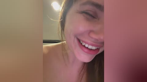 Video of a smiling woman with fair skin, brown hair, and bare shoulders, captured from a close-up angle, set against a blurred indoor background with a light source.