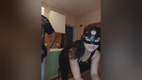 Video of a woman with long black hair and fair skin, wearing a black mask and revealing top, leaning over a door frame, revealing a bathroom with green cabinets and a white sink.