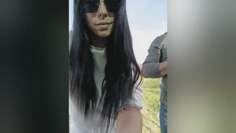 Video of a woman with long black hair, wearing sunglasses and a white t-shirt, standing outdoors on a sunny day with grassy background and blurred people in the distance.