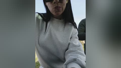 Video of a woman with long black hair, wearing sunglasses, a light grey sweater, and a slight pout. The background shows a blurred, outdoor scene with greenery and a clear blue sky.