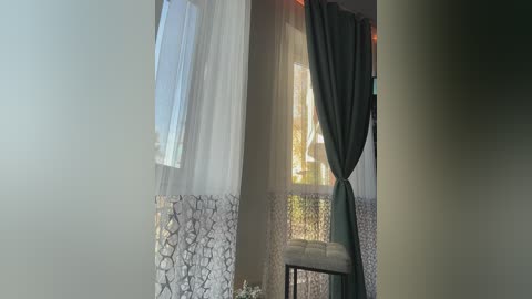 Video of a cozy room with a window dressed in sheer white curtains with intricate lace trim, teal drapes partially drawn, and a small cushioned stool.