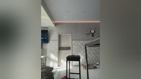 Video of a minimalist, modern bedroom with a light gray tiled wall, a flat-screen TV, a black stool, and a bed with a plush blanket.