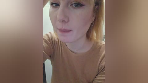 Video of a fair-skinned, light-haired woman with blue eyes, wearing a tan shirt, standing in a bathroom with blurry beige walls and a mirror reflection.