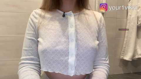 A video of a light-skinned woman with long brown hair wearing a sheer white lace crop top that reveals her nipples and cleavage. Background includes beige tiled walls and a towel hanging on a rack.