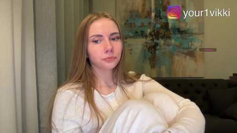 Video of a young Caucasian woman with fair skin, long blonde hair, and a neutral expression, wearing a white hoodie, sitting on a dark couch in a modern living room with abstract art on the wall.