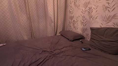Video of a dimly lit bedroom with a bed covered in brown bedding, a pillow, and a black smartphone on the mattress. The wallpaper features a light beige pattern.