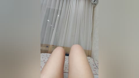 Video of a person's bare, light-skinned legs lying on a bed with white floral sheets, a white curtain covering a window, and an air conditioner on the wall in a simple, softly lit bedroom.