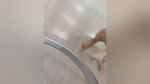 A close-up video of a person's hand, with a blurred, light-colored wooden floor and a metallic object in the background. The image emphasizes textures and a minimalist aesthetic.