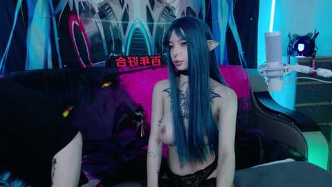 Video of a topless, pale-skinned Asian woman with blue hair and pointed ears, wearing black lace lingerie, sitting on a pink couch with a black cat next to her in a dimly-lit room.