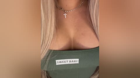 A video of a woman with long blonde hair, wearing a green ribbed top with \"SWEET BABY\" text, showcasing her ample cleavage and a silver necklace with a cross pendant.