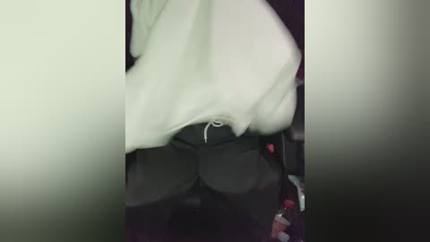 A dimly lit video shows a person with a light skin tone, wearing a white hoodie, black pants, and a visible zipper on their pants, holding a bottle. The background is blurry and dark.