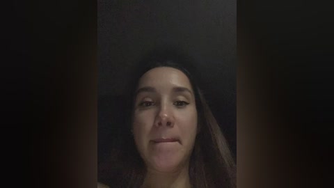 A video of a young woman with long, dark hair, light brown skin, and a neutral expression, set against a dark background.