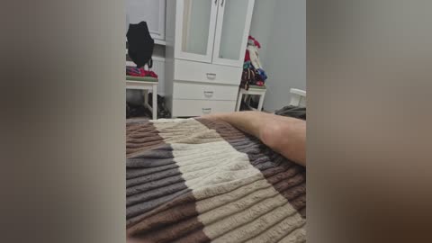 A video of a cozy bedroom featuring a large, patterned knit blanket with brown, beige, and white stripes. White cabinets and a white dresser with a black jacket on top are in the background.
