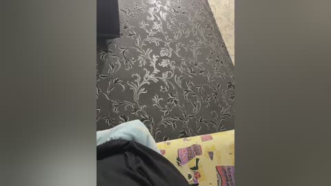 A video shows a dark gray carpet with a floral pattern, a black chair, and a yellow sheet with cartoon characters. The image is slightly blurry, possibly taken with a shaky camera.